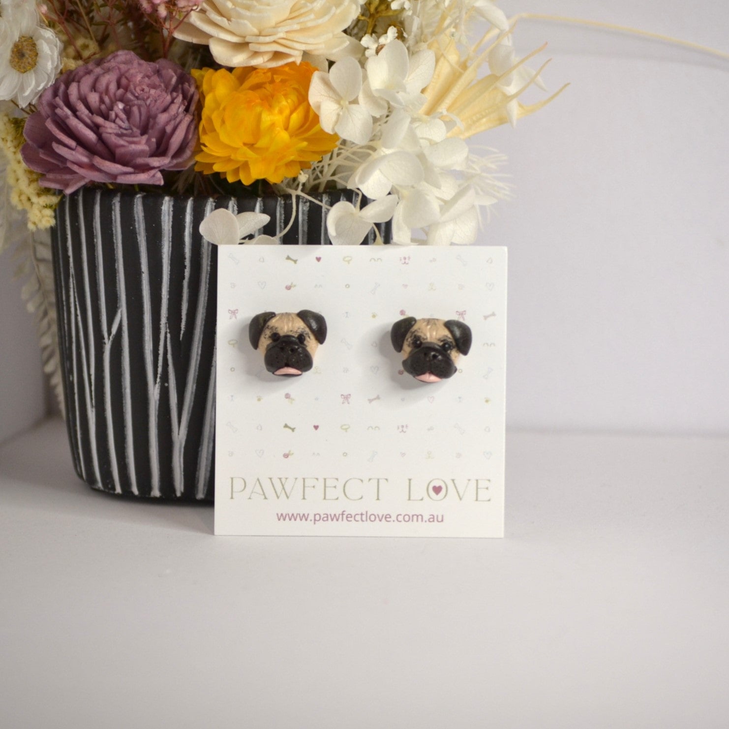 Handmade Pug stud earrings by Pawfect Love, positioned in front of dried flower arrangement