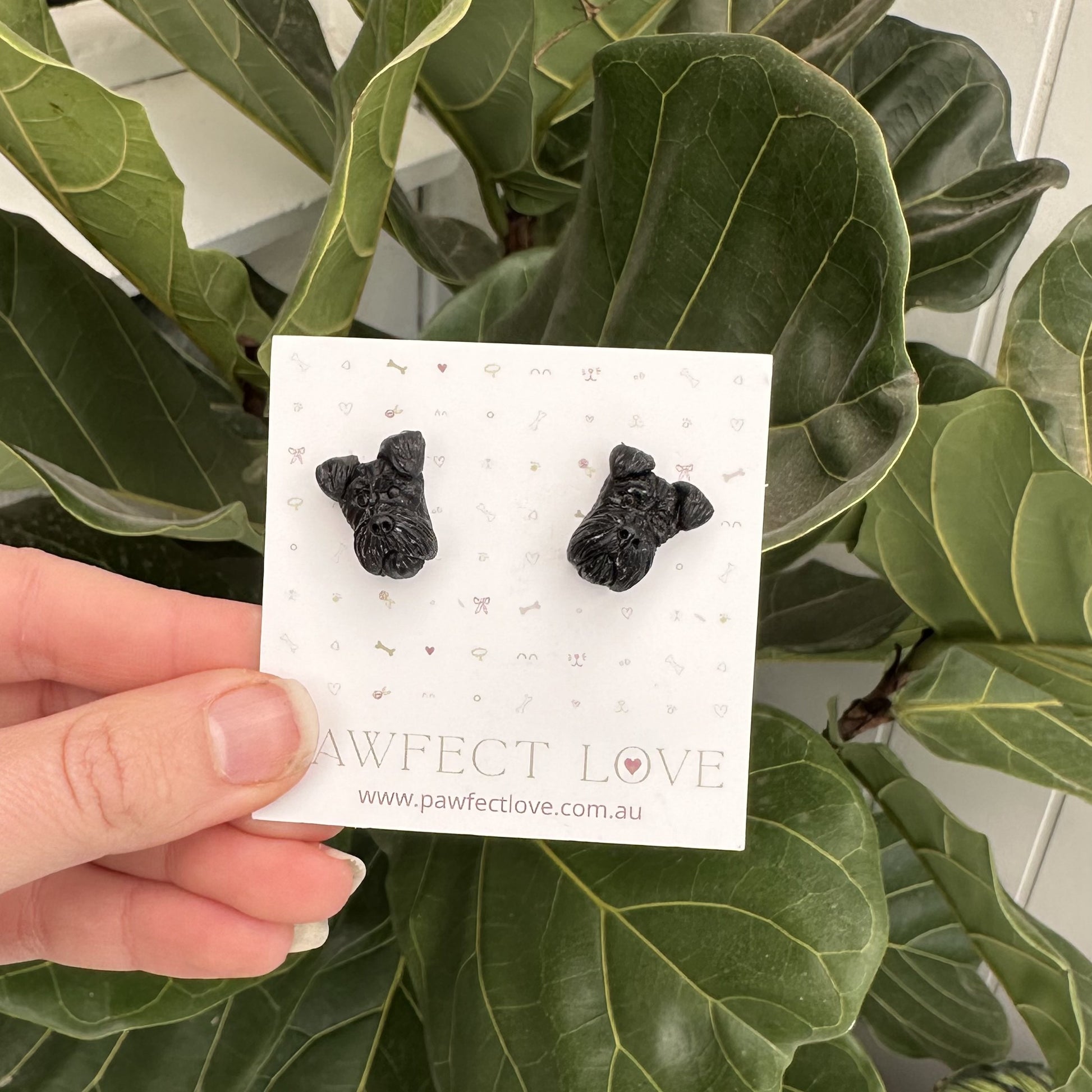 Black schnauzer dog-shaped earrings on a card held in front of green leaves