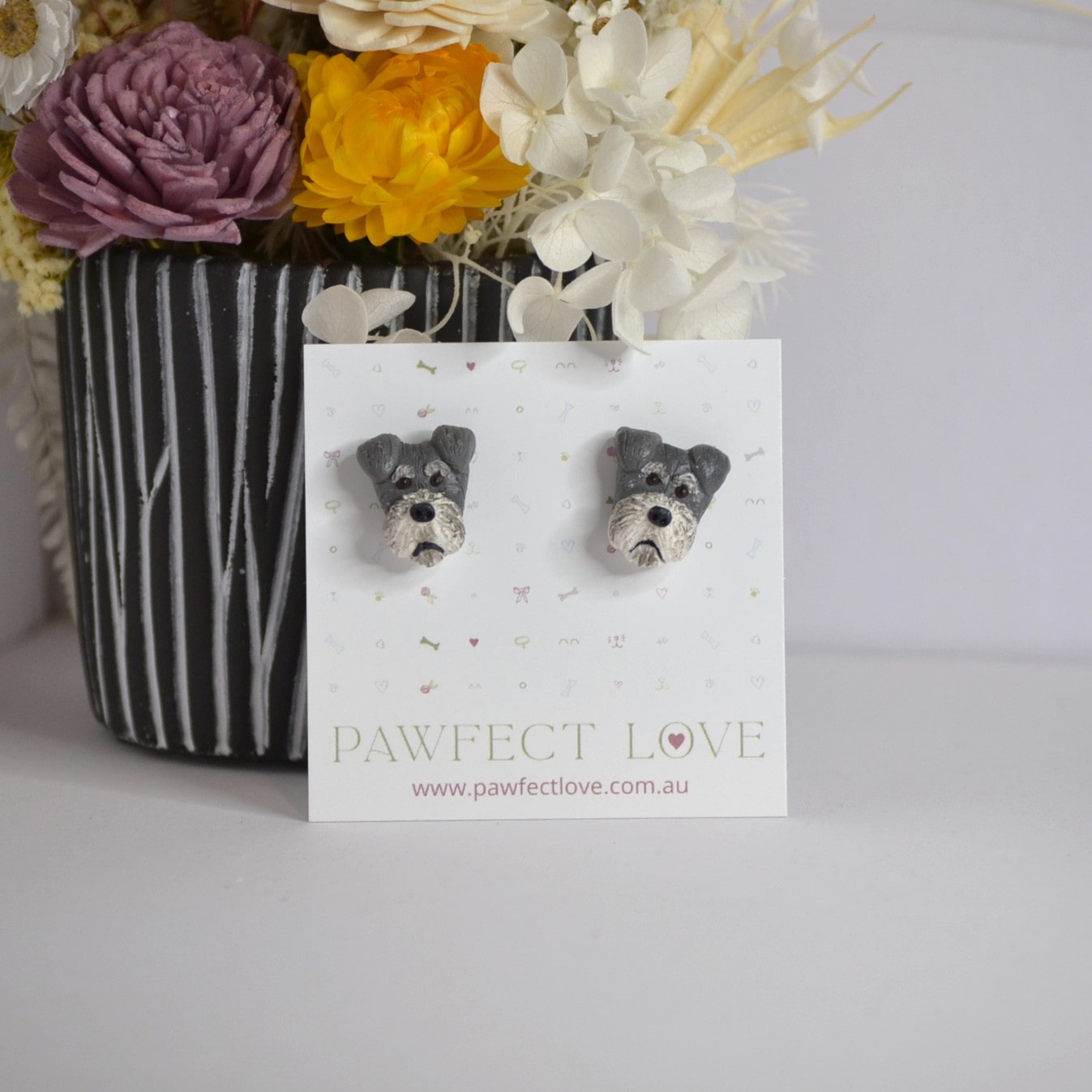 Handmade schnauzer stud earrings by Pawfect Love, positioned in front of dried flower arrangement