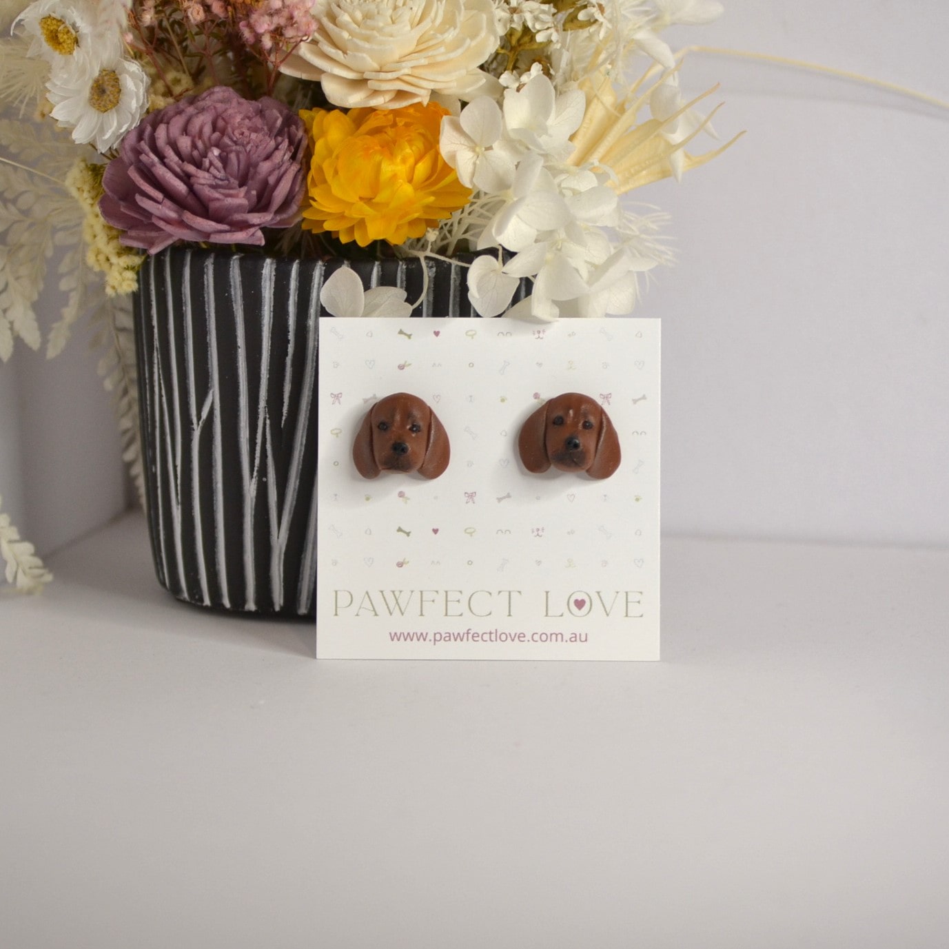 Handmade dachshund stud earrings by Pawfect Love, positioned in front of dried flower arrangement
