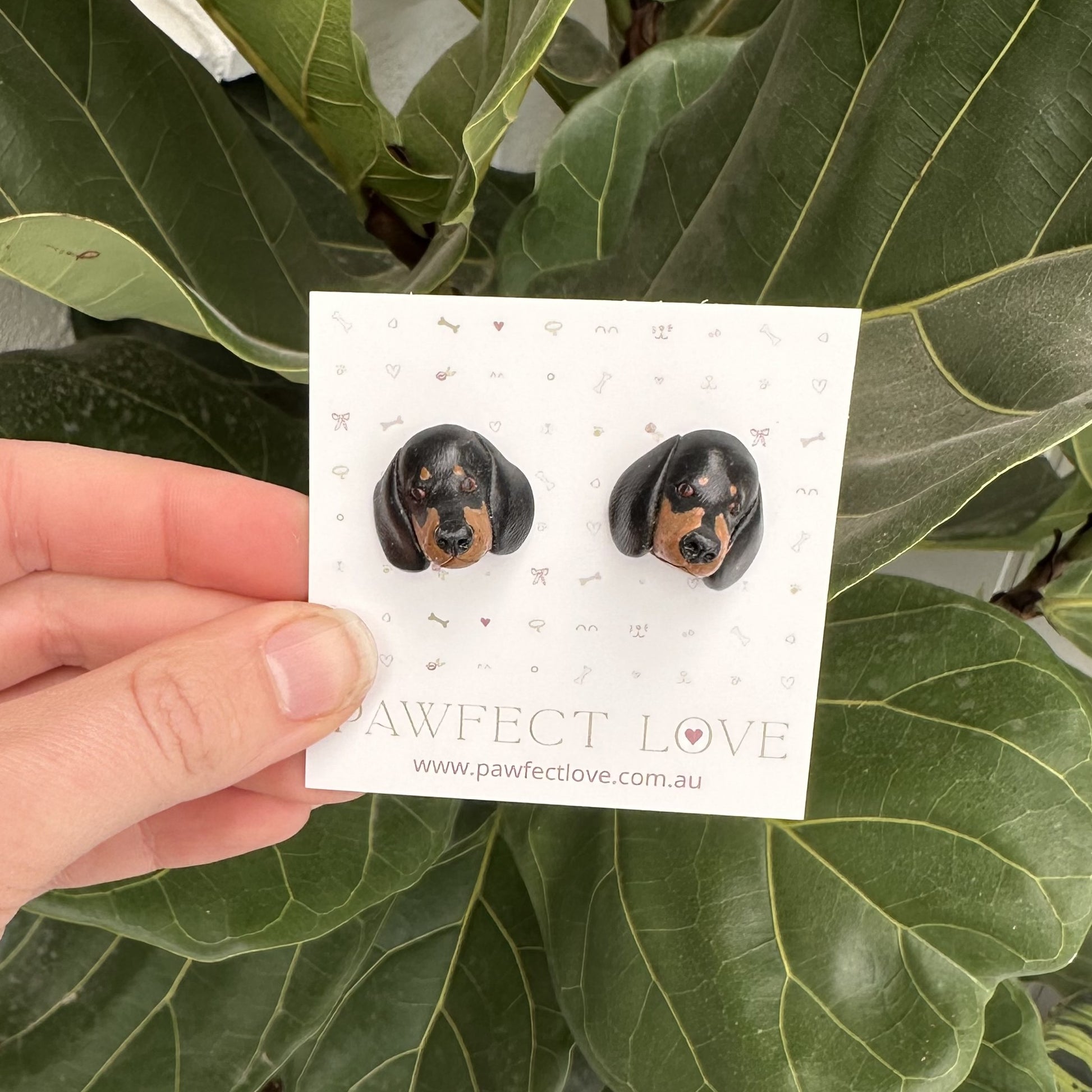 Pair of black and tan dachshund dog-shaped earrings on a 'Pawfect Love' branded card held over green leaves.