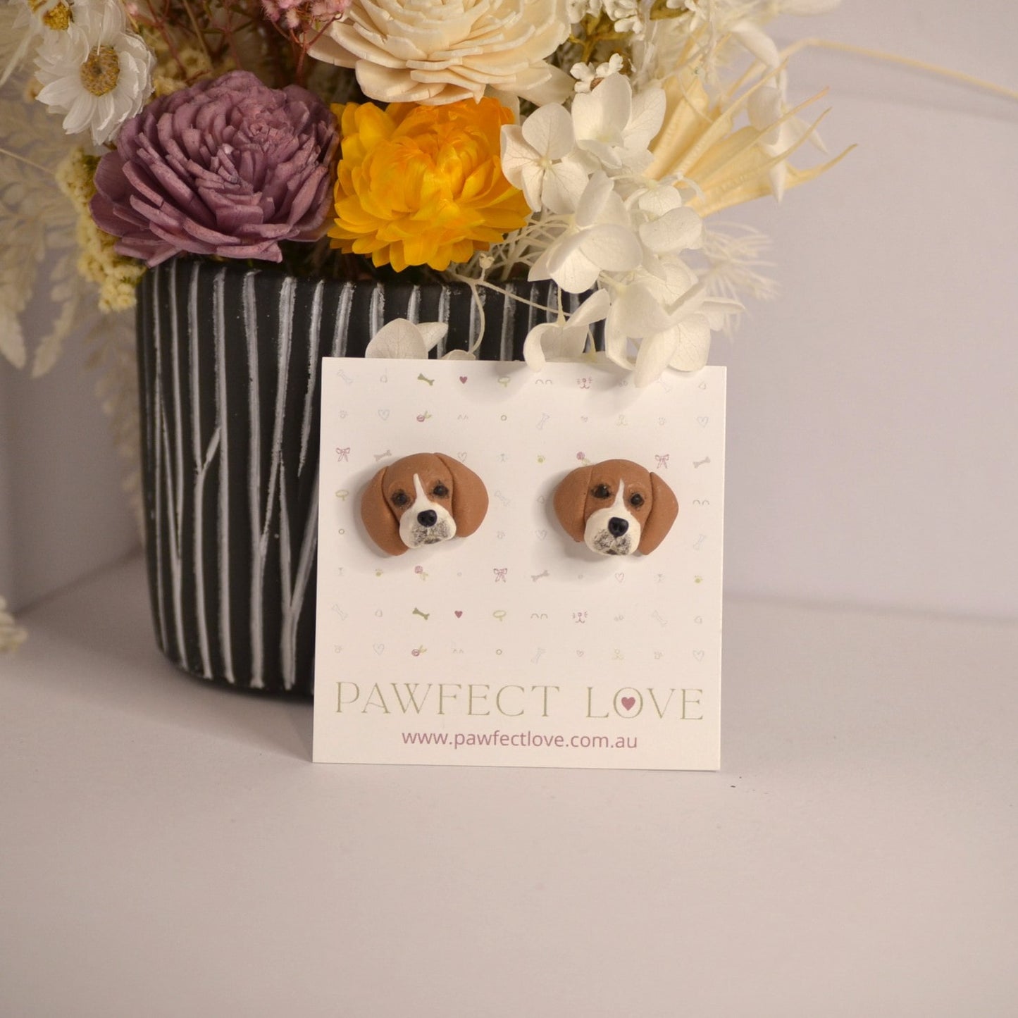 Handmade beagle stud earrings by Pawfect Love, positioned in front of dried flower arrangement