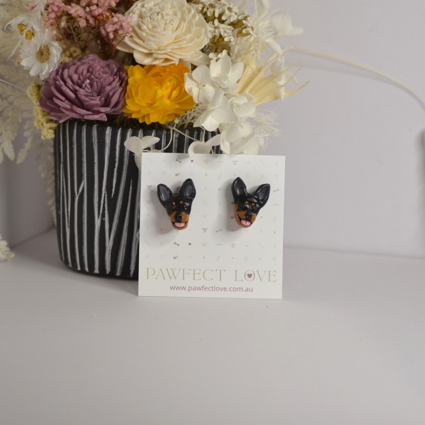 Handmade kelpie stud earrings by Pawfect Love, positioned in front of dried flower arrangement
