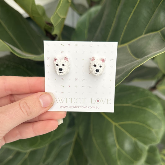 Hand holding a card with Westie dog-shaped earrings against a green leafy background
