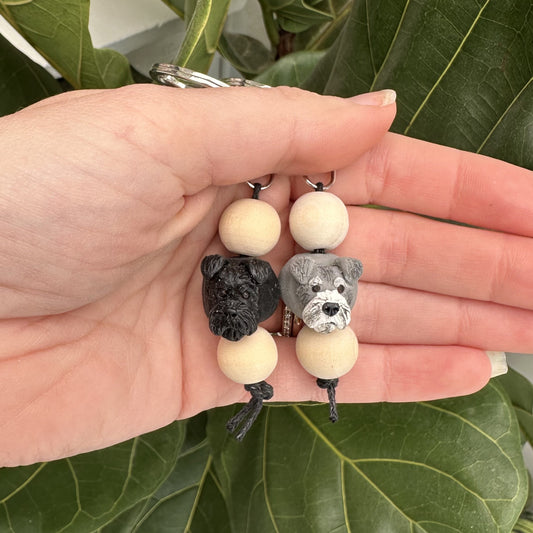 Hand holding two keychains with schnauzer dog-shaped beads against a leafy background