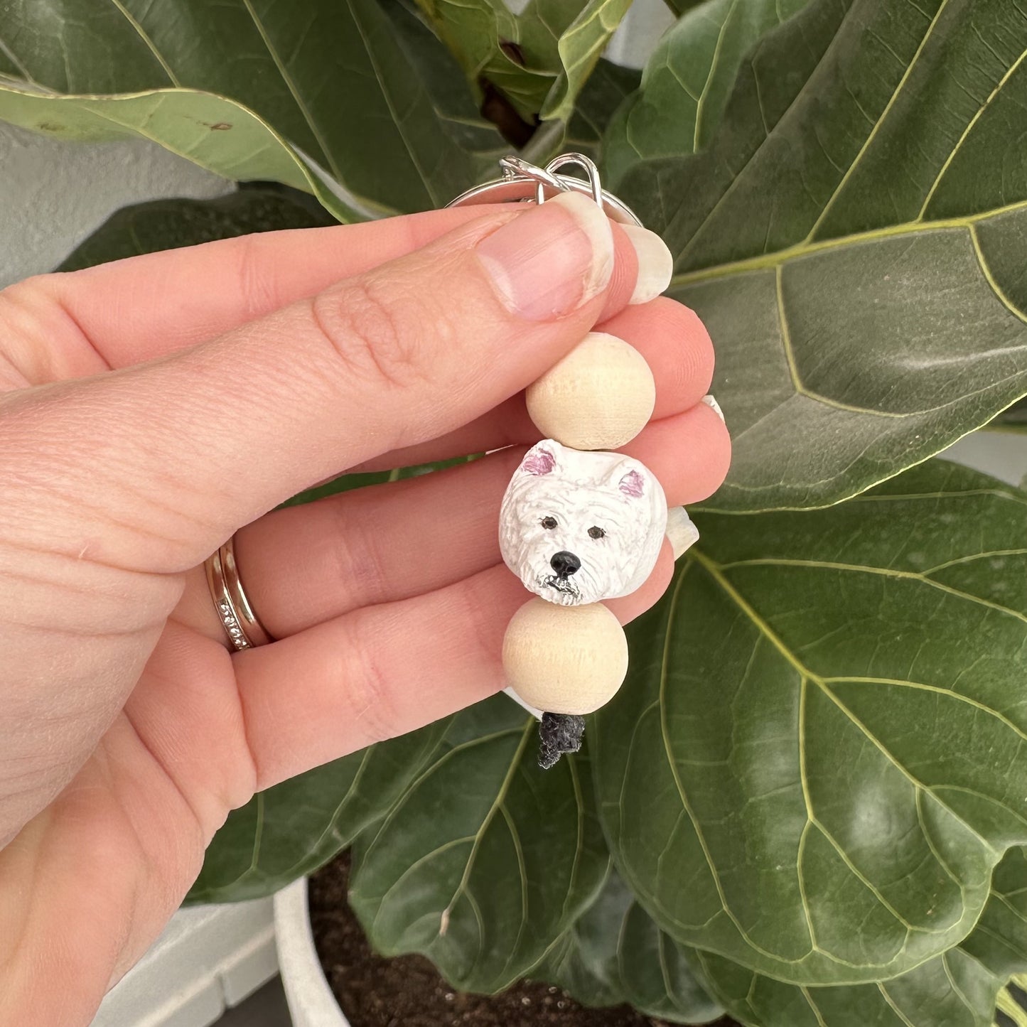 Hand holding a beaded Westie dog keyring with a dog design against a leafy background