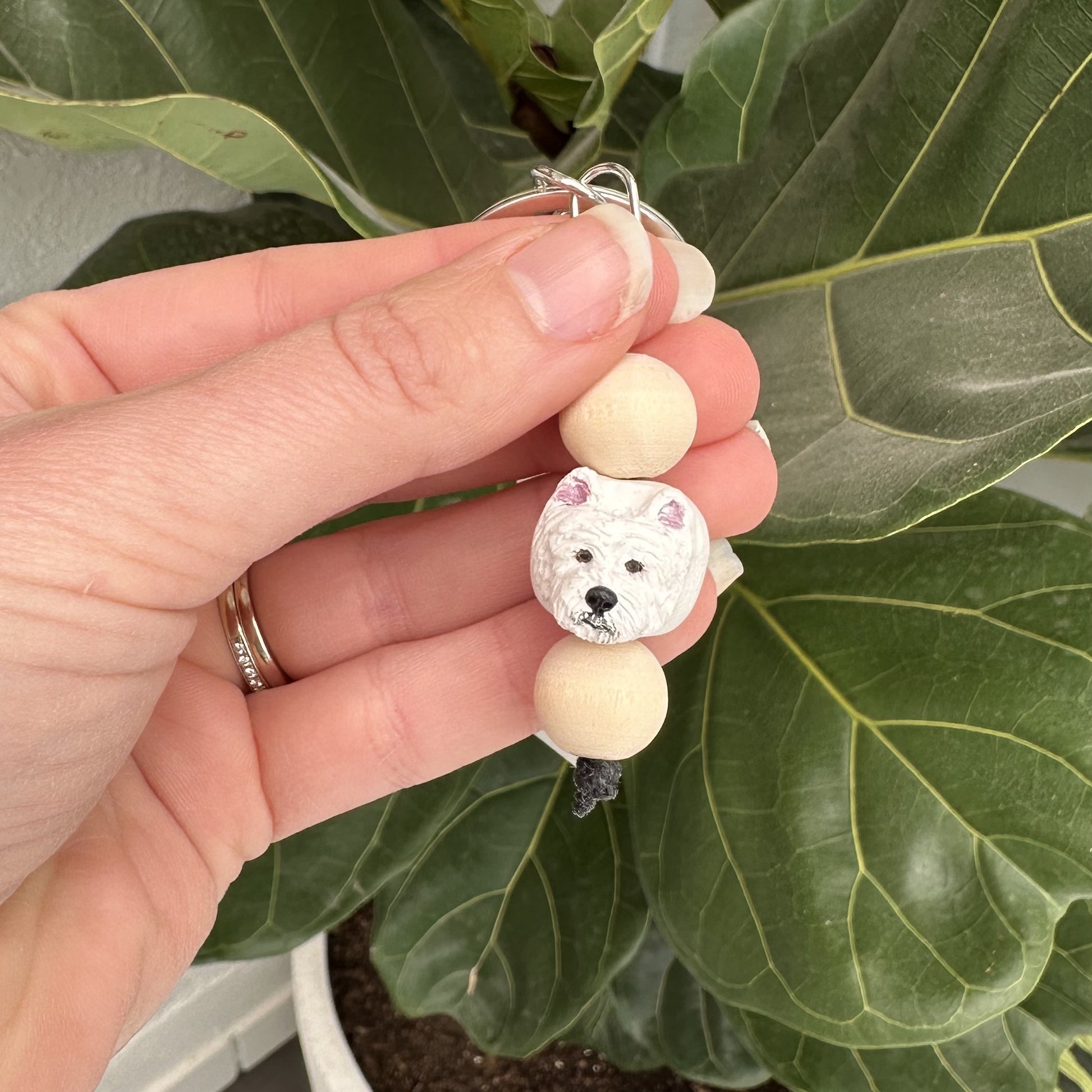Hand holding a beaded Westie dog keyring with a dog design against a leafy background