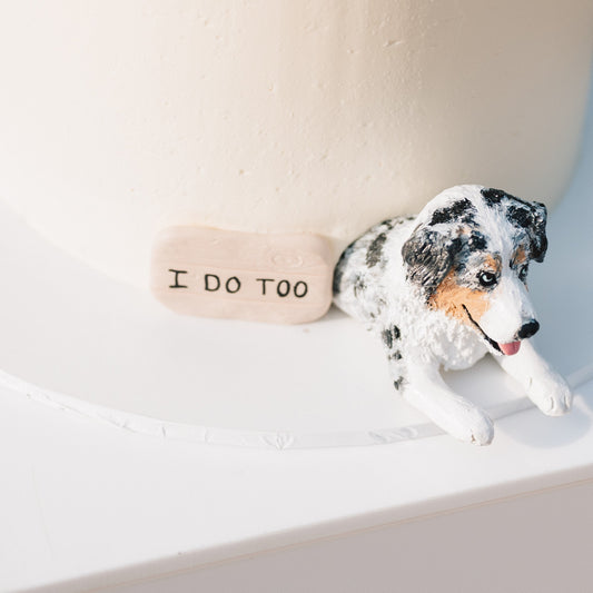 Handmade custom pet cake topper of an Australian Shepherd cake topper peeking out of a wedding cake, beside a sign that says 'I DO TOO'.