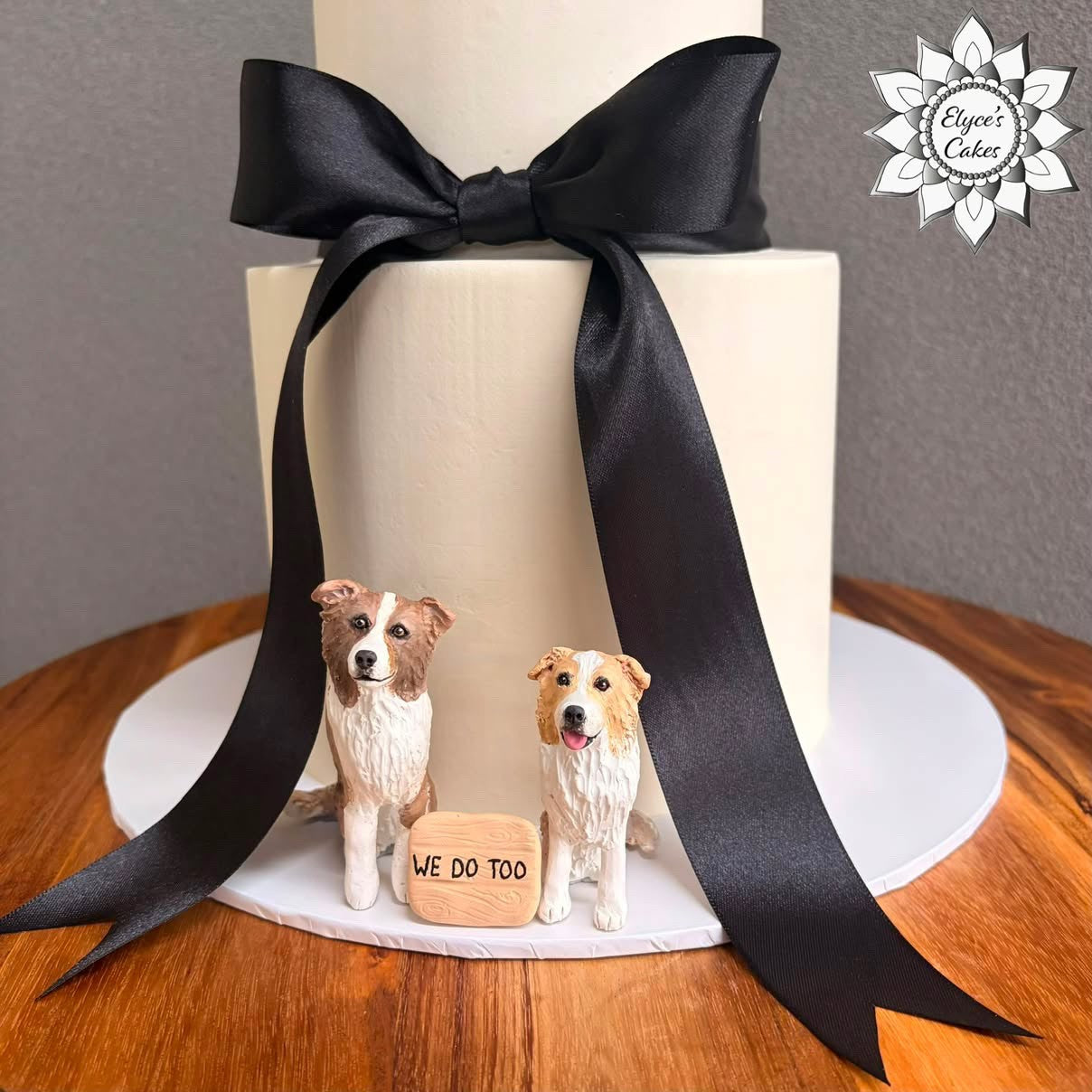 A custom two-tiered cream cake decorated with a large black satin bow and two small dog figurines standing beside a wooden sign that reads 'WE DO TOO'. The cake sits on a wooden table against a neutral gray background, with a 'Elvia's Cakes' logo in the top right corner.