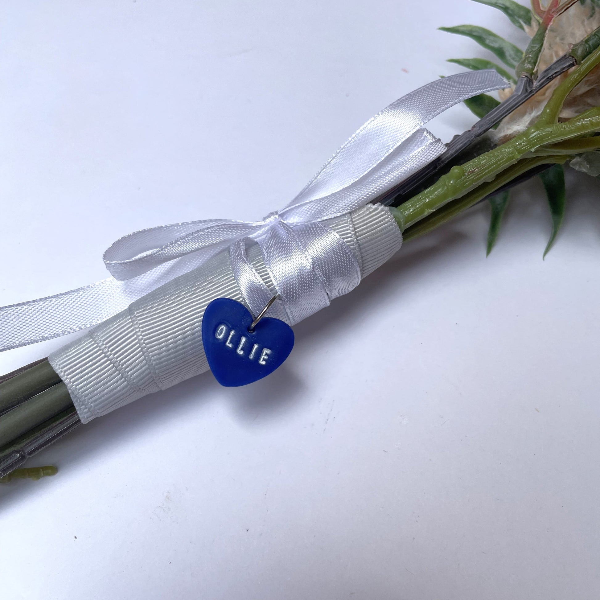 Blue heart bouquet accessory on ribbon around stems, with white name 'Ollie' imprinted in white.