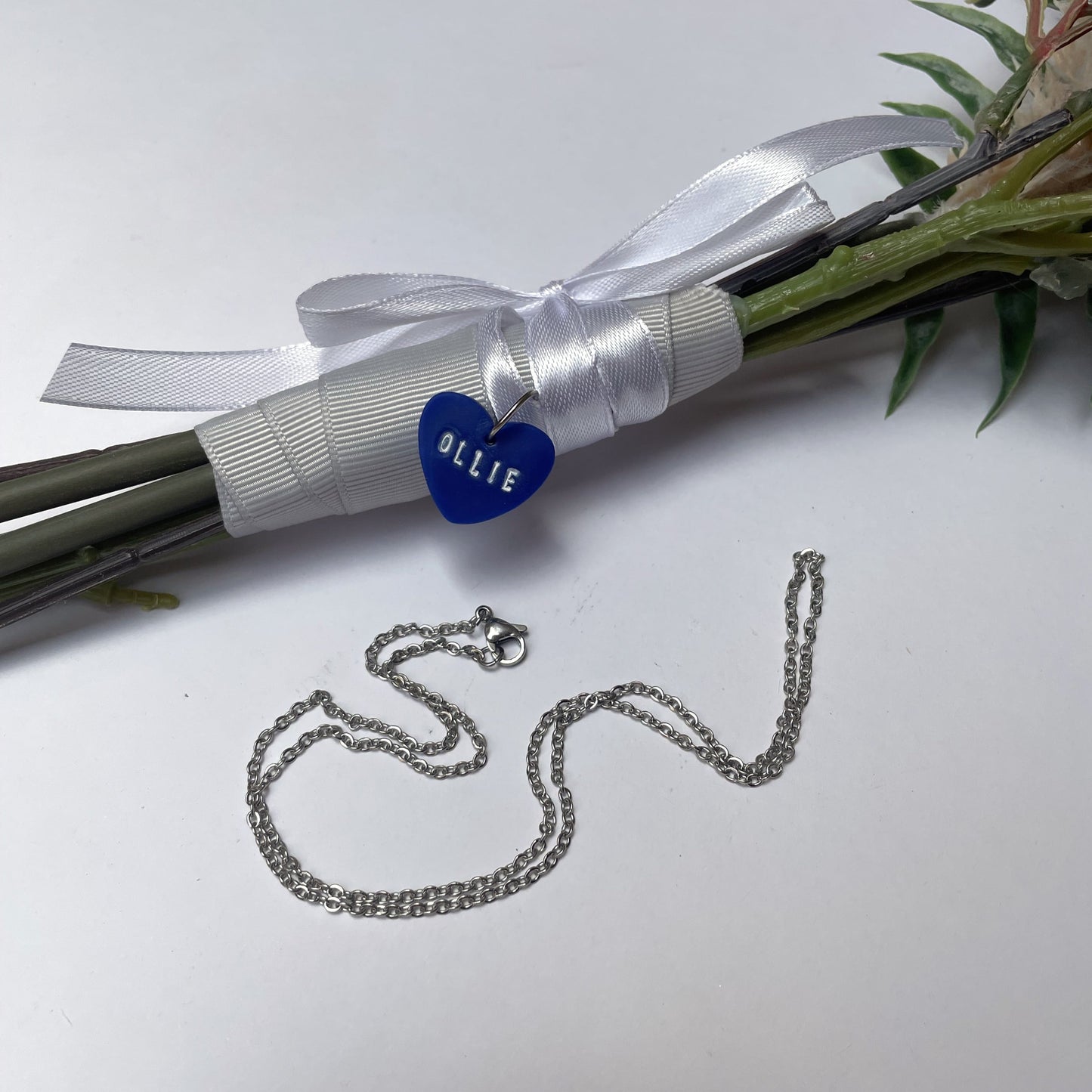 Blue heart handmade bouquet pendant with name 'ollie' in white. In the foreground is a silver necklace chain.