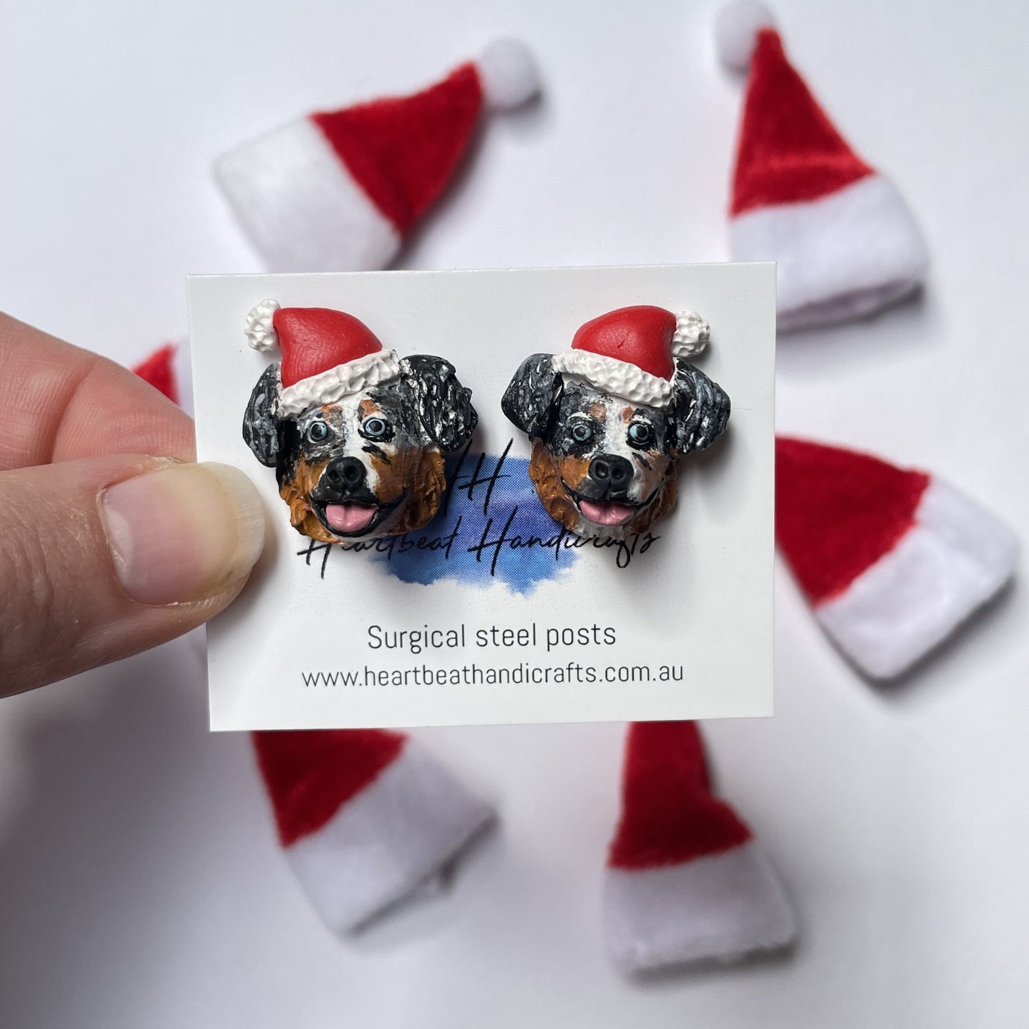 Handmade earrings of a Australian Shepherd dog wearing santa hats, made from polymer clay held over a Christmas hat background.