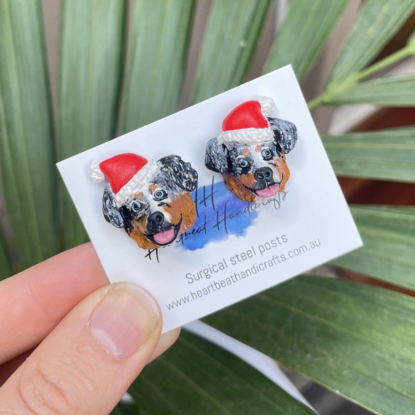 Handmade earrings of a Australian Shepherd dog wearing santa hats, made from polymer clay, held over a green palm tree background.