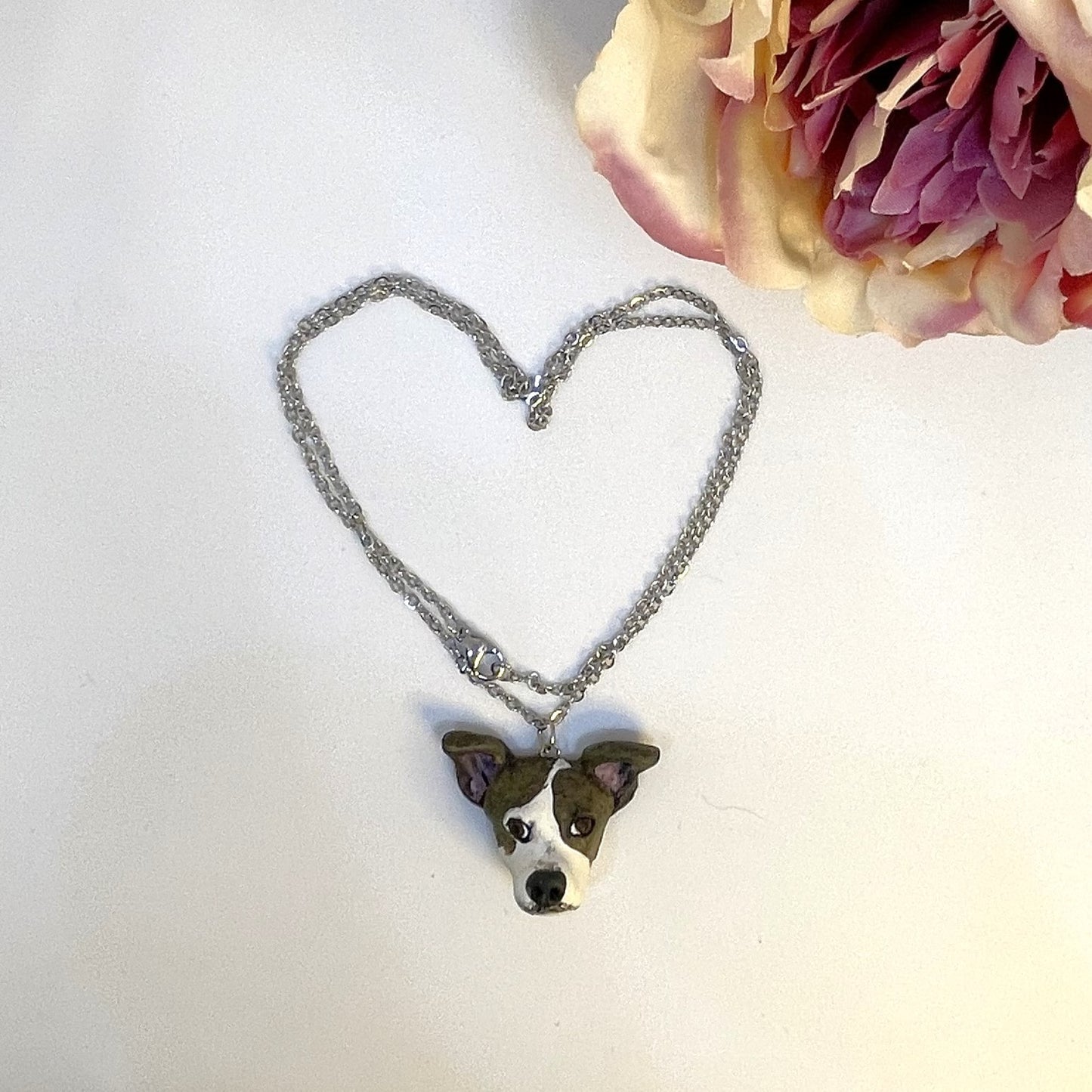 Handmade custom brown and white dog necklace pendant on chain arranged in a heart shape, in front of faux pink flower.