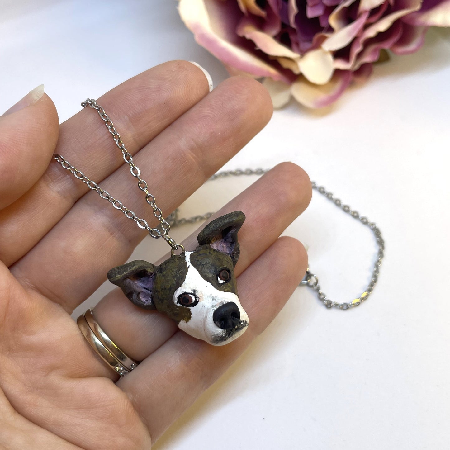 Handmade custom brown and white dog necklace pendant on chain held in front of faux pink flower.