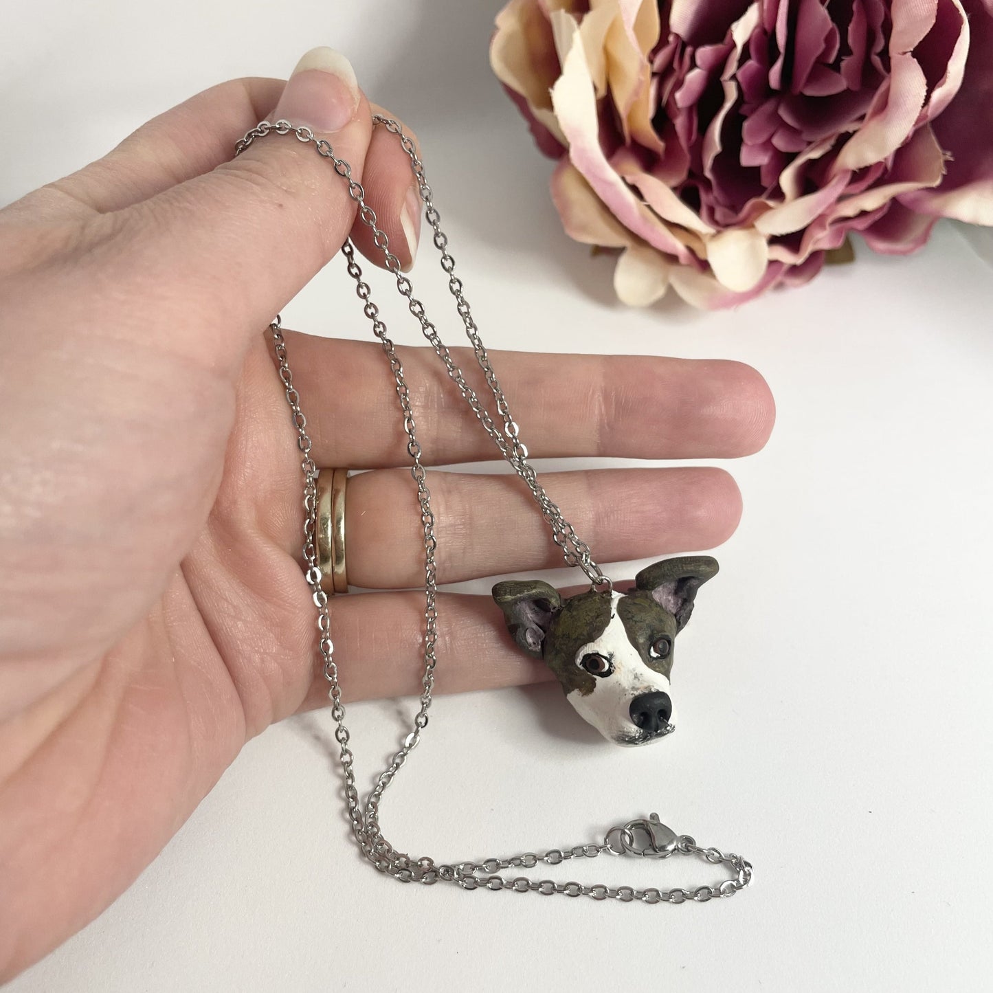 Handmade custom dog face necklace pendant on chain, held in front of faux pink flower.