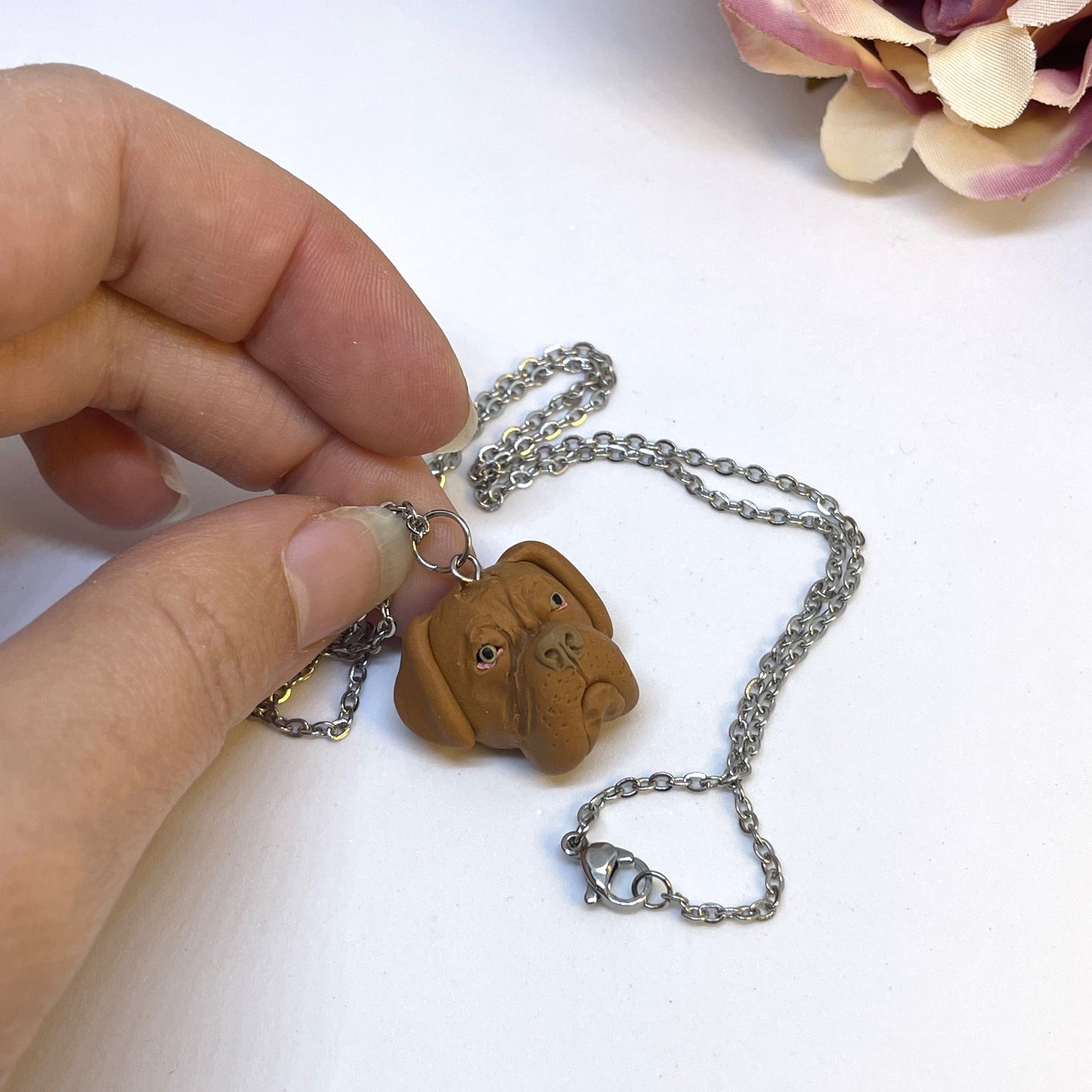 Handmade custom dogue de bordeaux face necklace pendant on chain, held in front of faux pink flower.
