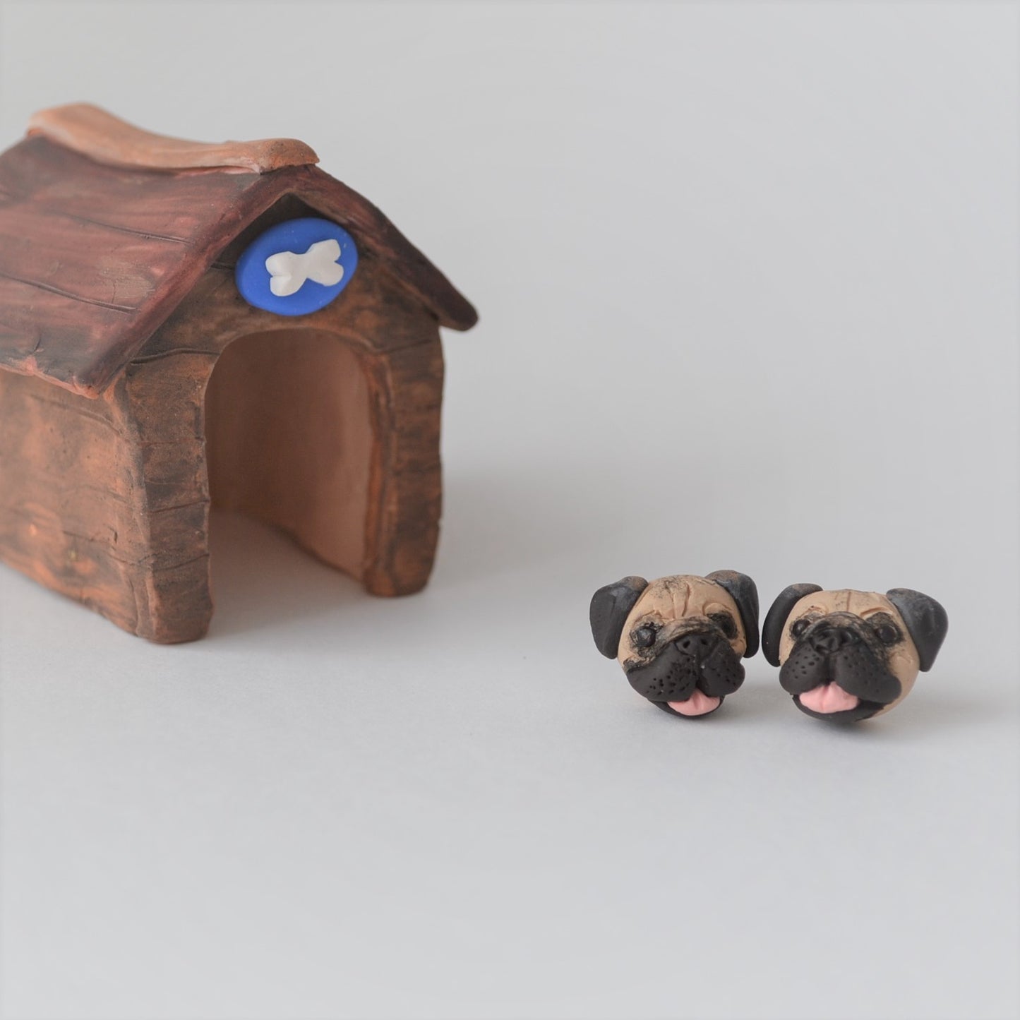 Handmade polymer clay pug stud earrings shown styled in front of a mini kennel
