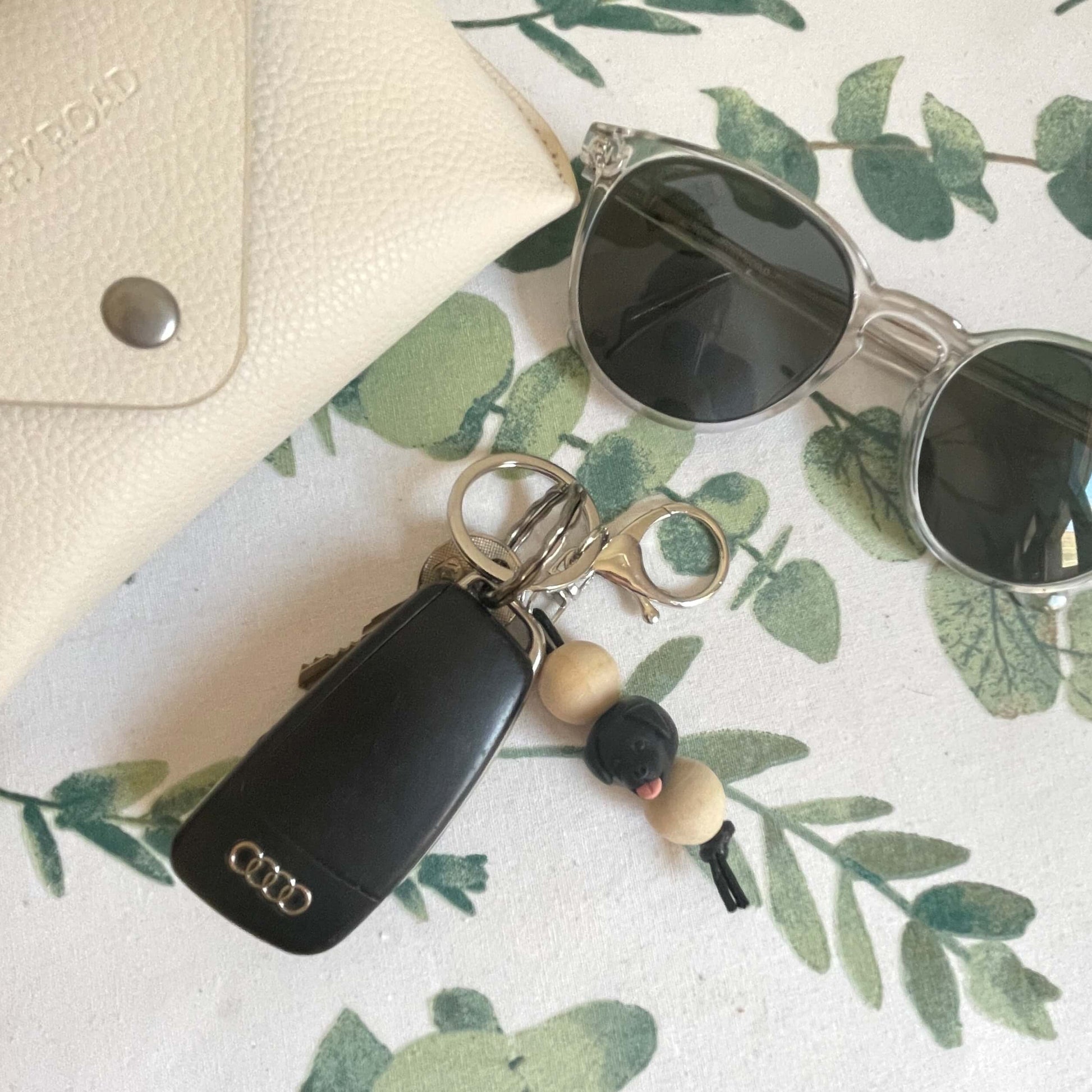 Handmade black labrador timber and polymer clay keychain laying on top of a white tablecloth beside sunglasses and a case