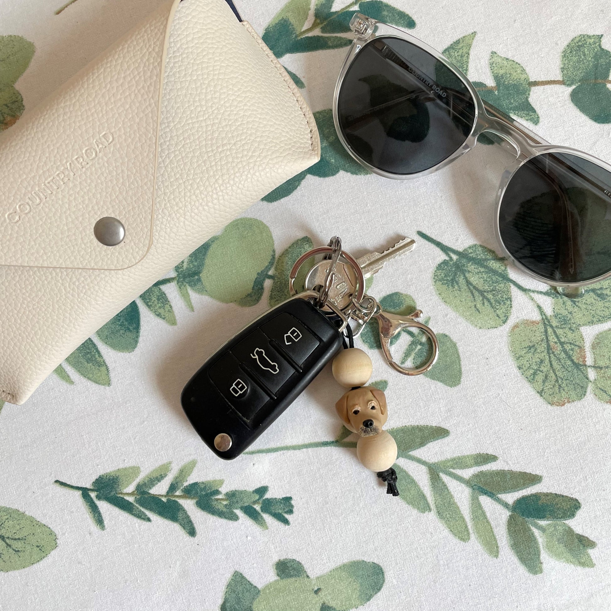 Handmade timber and polymer clay golden retriever dog keychain on white floral tablecloth beside sunglasses