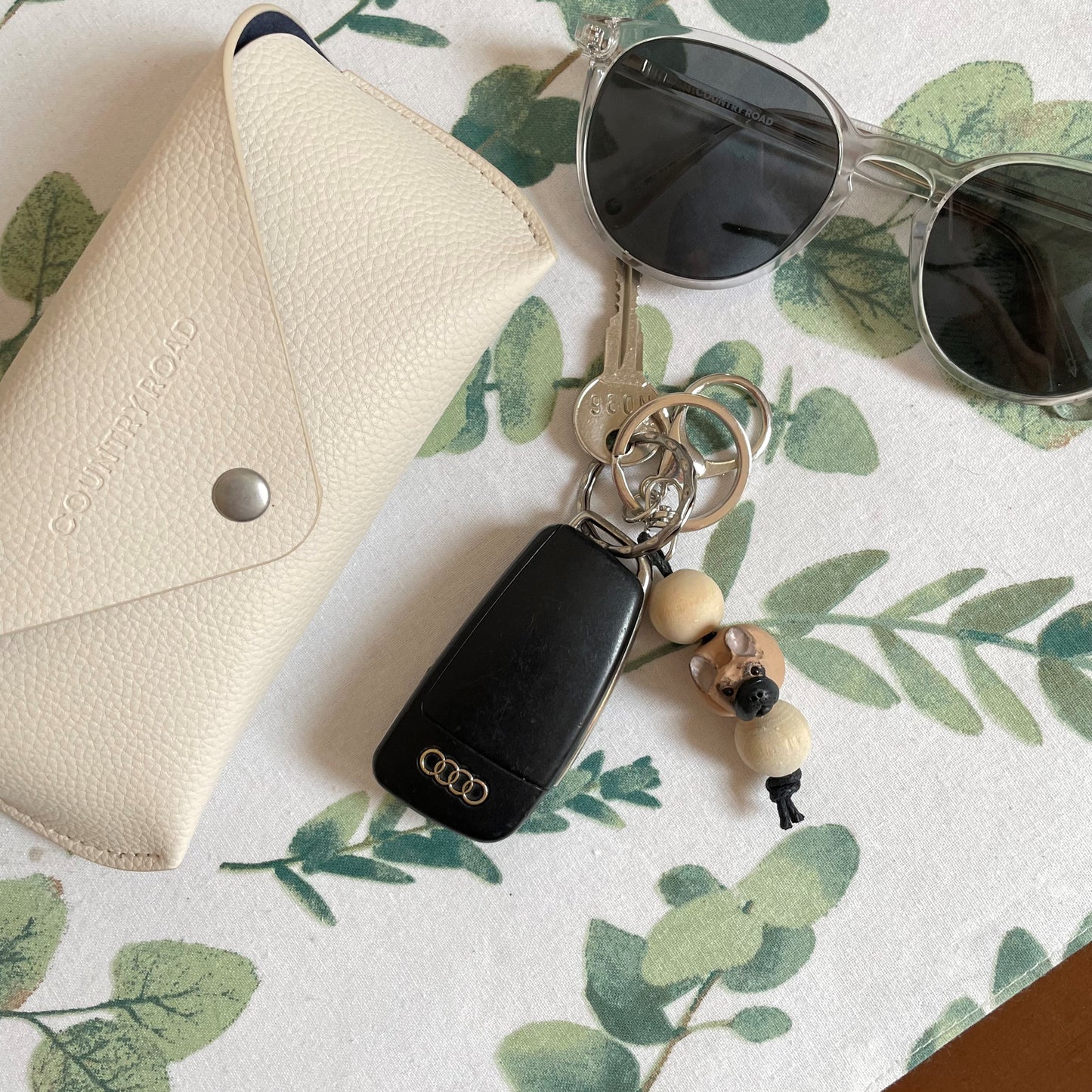 Handmade timber and polymer clay french bulldog keychain on white tablecloth beside sunglasses