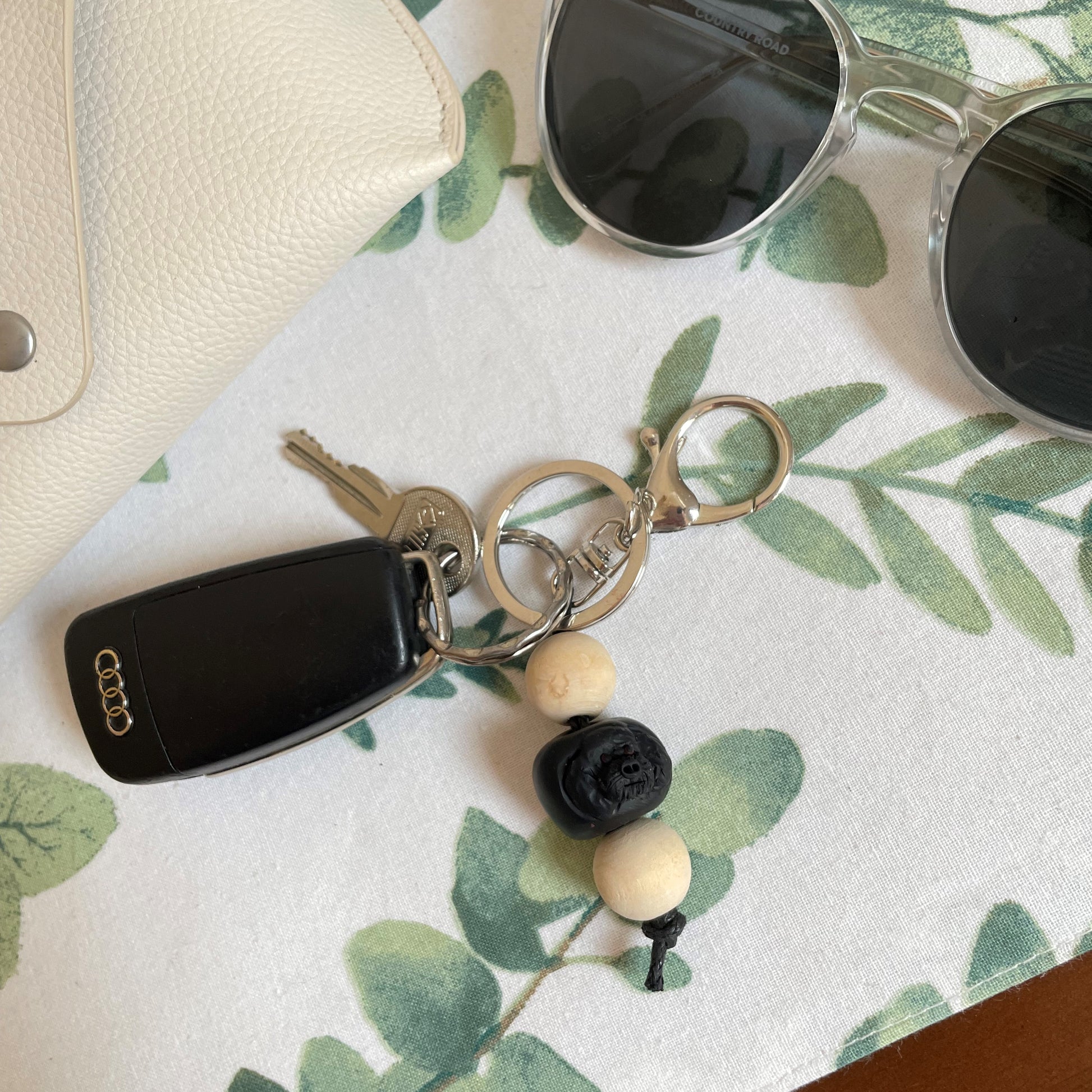 Handmade timber and polymer clay black poodle keychain on a tablecloth beside sunglasses and a case.