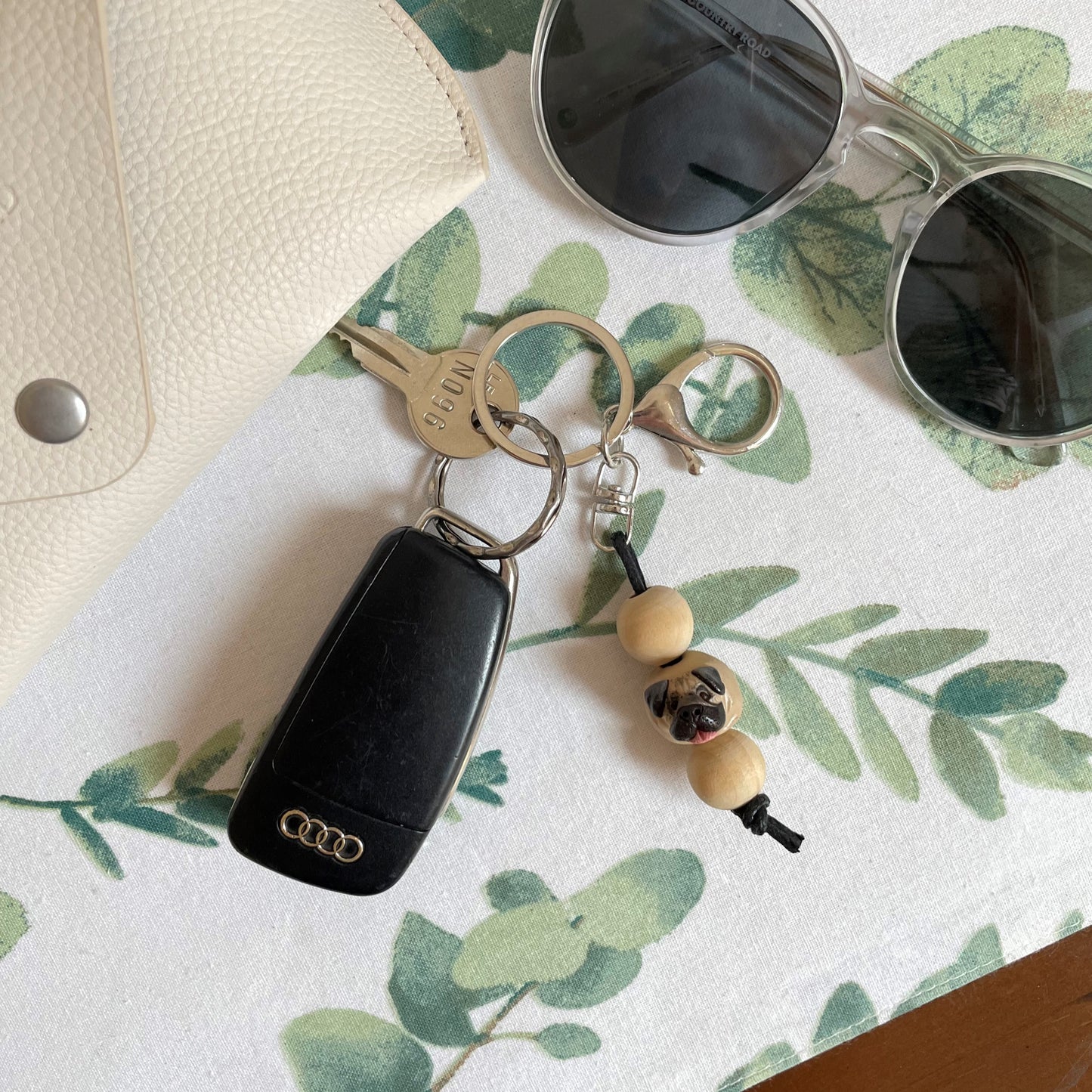 Handmade timber and polymer clay pug dog keychain on white floral tablecloth beside sunglasses