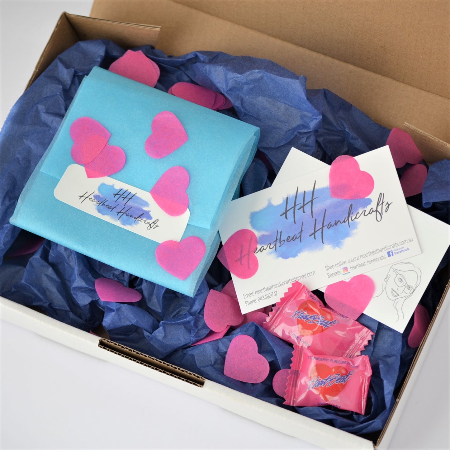 Example of packaging of order, showing blue tissue paper wrapping, pink heart-shaped confetti and a Heartbeat Handicrafts business card